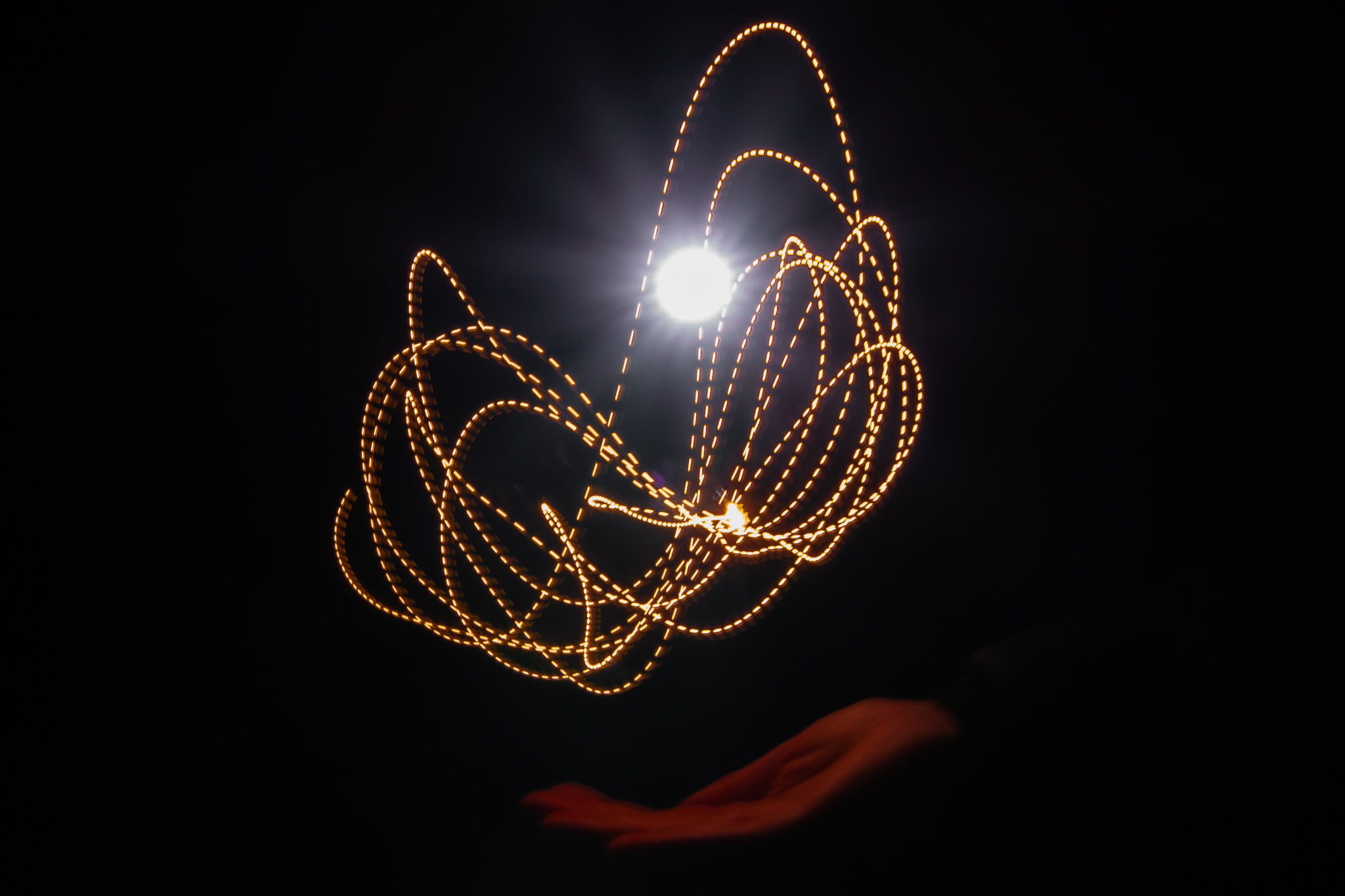 A bright white orb of light and dotted curvy orange light trails hover above a hand lit with orange light