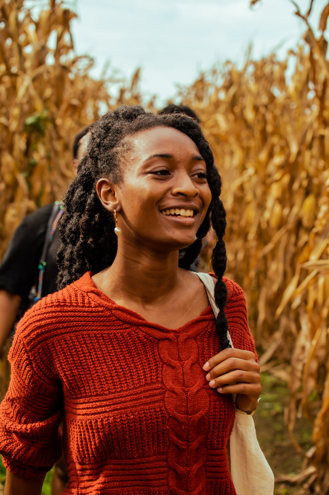 a young adult walks through a corn maze smiling with a bag slung across their shoulder